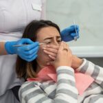 mloda-kobieta-siedzaca-w-klinice-dentystycznej-na-krzesle-z-atakiem-paniki-zakrywajac-usta-obiema-rekami-jej-koszmarna-procedura-stomatologiczna